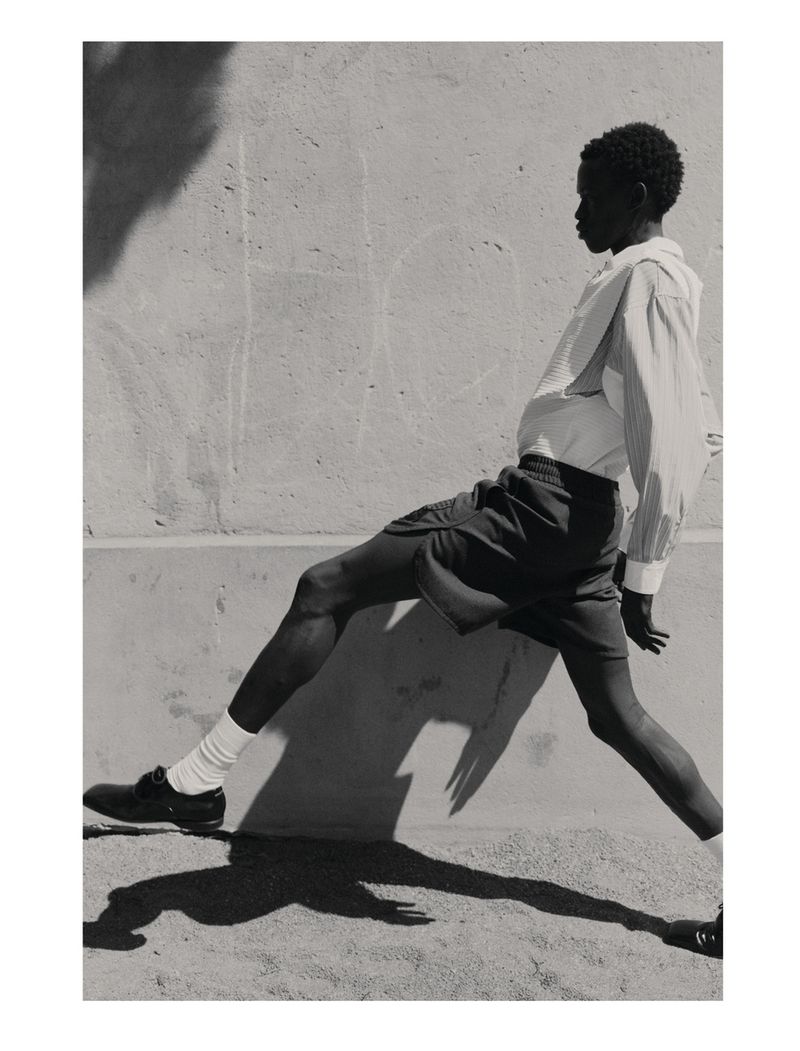 Monochrome image of a slender, dark-skinned adult striding, legs wide, captured in bright sunlight against a neutral wall backdrop.