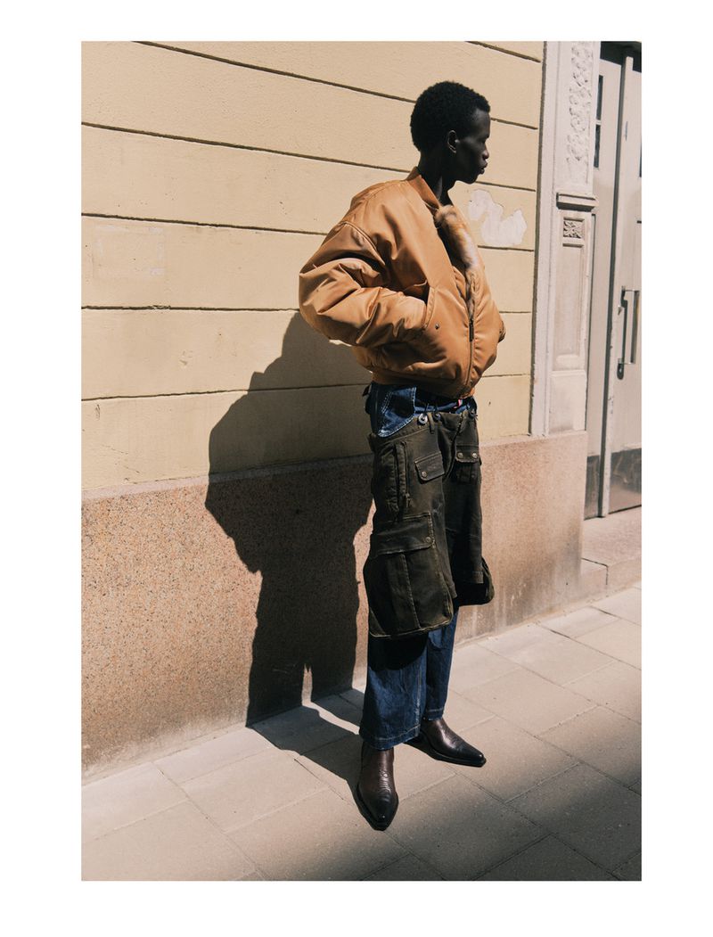 An African American male model is showcased in an outdoor setting wearing a bomber jacket, cargo pants, and boots in a fashion editorial.