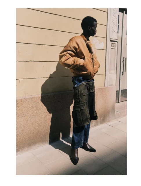 An African American male model is showcased in an outdoor setting wearing a bomber jacket, cargo pants, and boots in a fashion editorial.