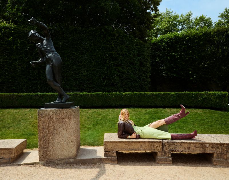 A woman in a brown jacket and green skirt is laying on a stone bench in front of a bronze statue in a park setting with lush green trees.