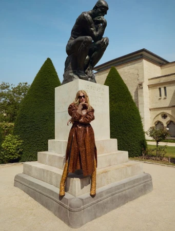 Woman in a snakeskin-print coat poses in front of Rodin's The Thinker sculpture in an outdoor garden setting with buildings.