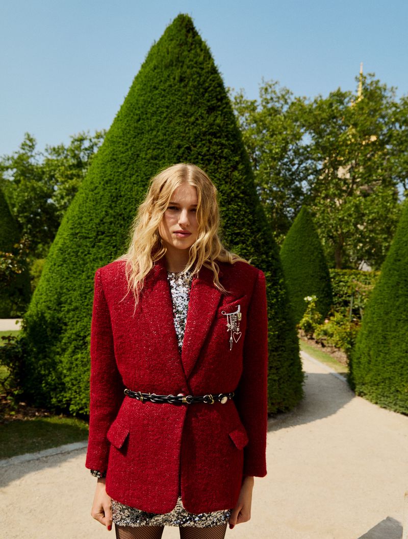 Fashionable woman poses in garden wearing red tweed blazer, sequin dress, and black belt. Green topiary and blue sky in the background.
