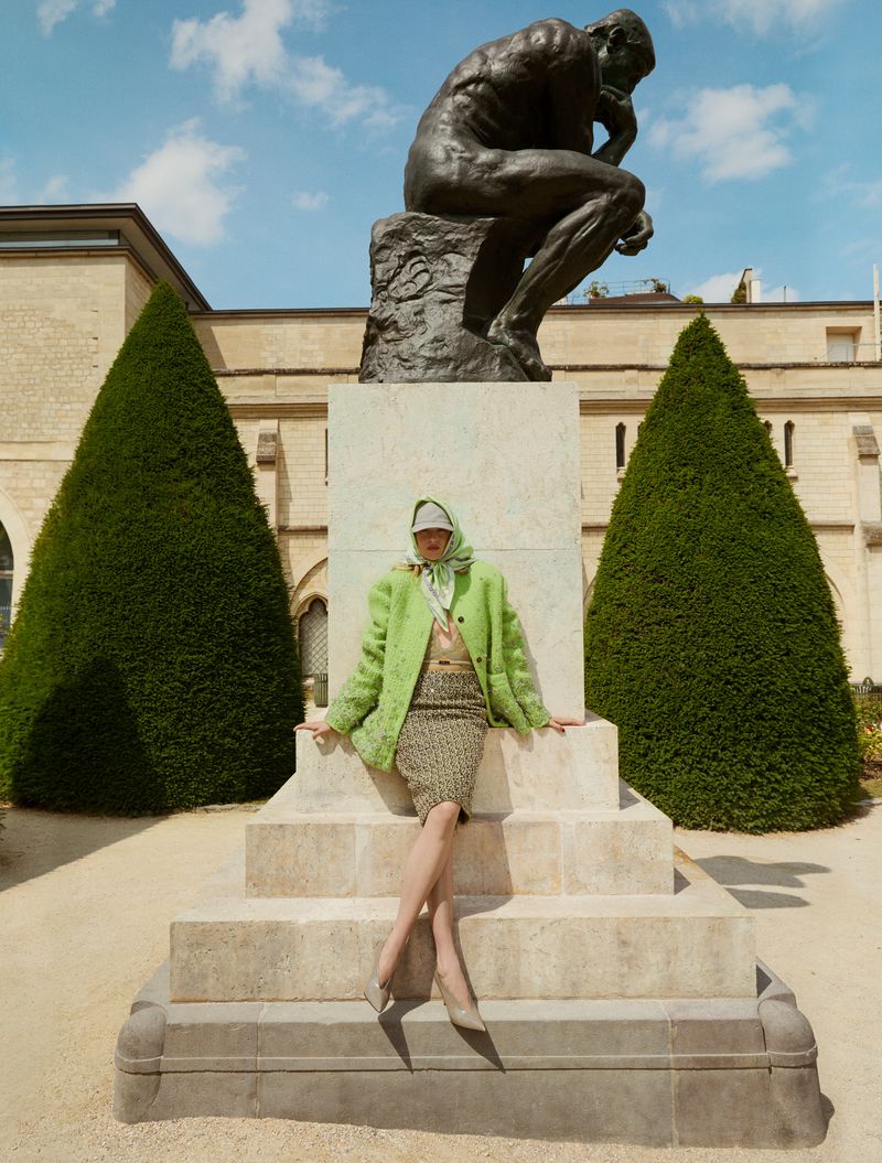 Fashion shot of a woman in a green jacket sitting in front of Rodin's 'The Thinker' statue in an outdoor setting with manicured landscaping.