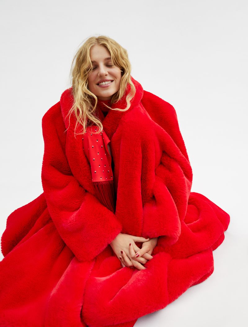 Woman in a red faux fur coat sitting on a white background with her eyes closed and hands clasped. Red manicure is visible.