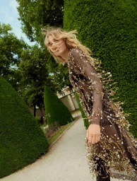 Model Dana Elmers wears a sheer black dress with gold sequin branch details along a path bordered by conical topiary hedges.