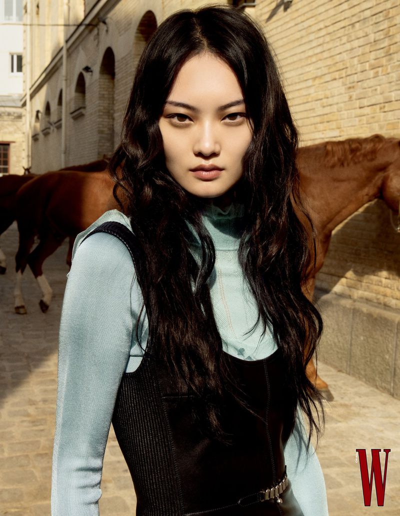 Portrait of an Asian woman with dark wavy hair wearing a dark dress with horses in the background on a cobblestone street.