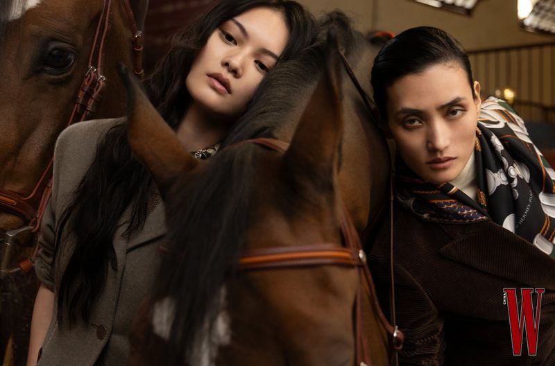 Two adult females pose in front of brown horses in an indoor stable, wearing brown and tan jackets, in a medium shot with soft lighting.