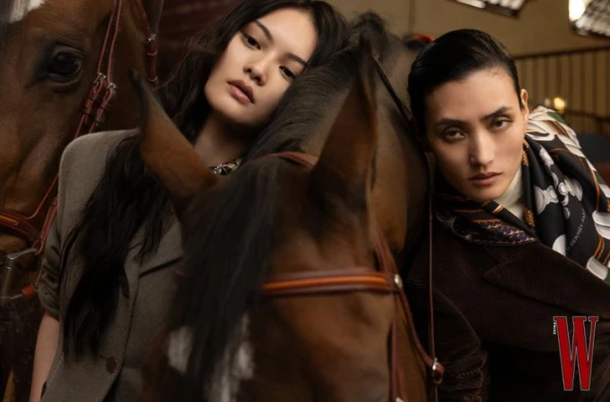 Two adult females pose in front of brown horses in an indoor stable, wearing brown and tan jackets, in a medium shot with soft lighting.