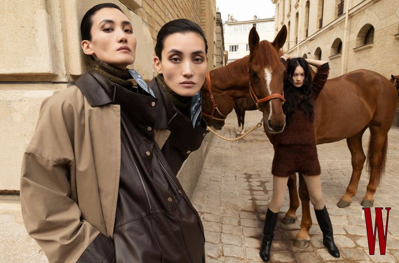 Two women in coats pose next to a brown horse on a cobblestone street with light-colored buildings in the background for a fashion editorial.