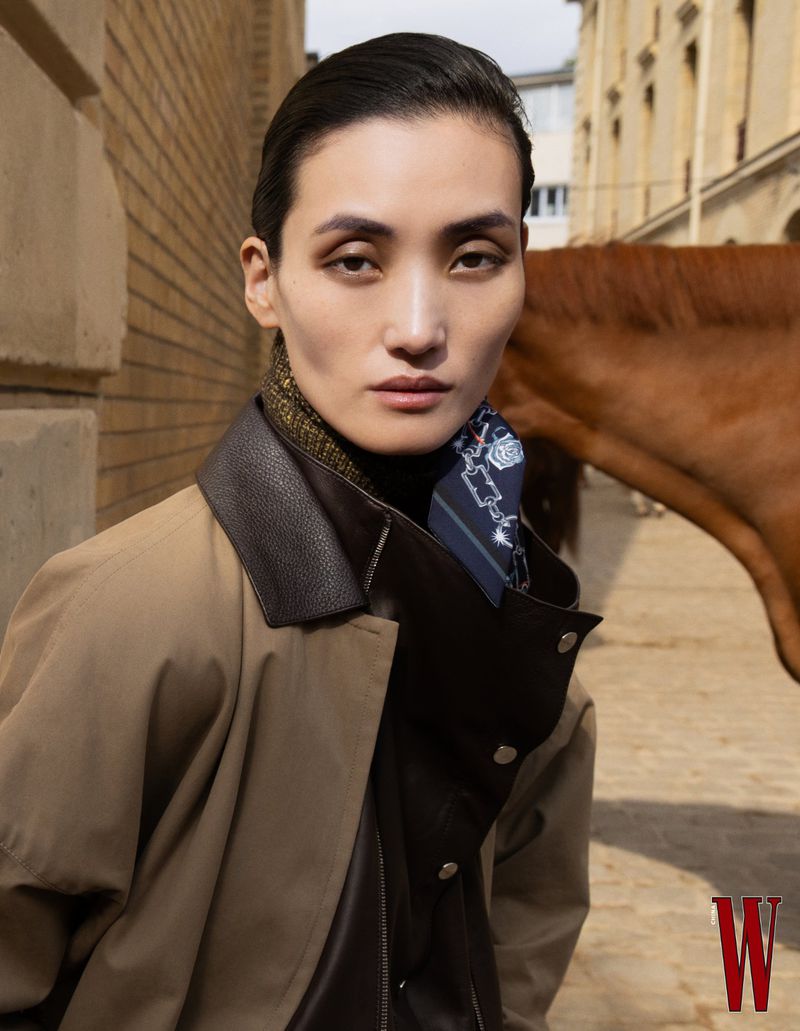 Asian woman poses outdoors wearing a tan trench coat, leather vest, and patterned scarf, with a horse visible in the background.