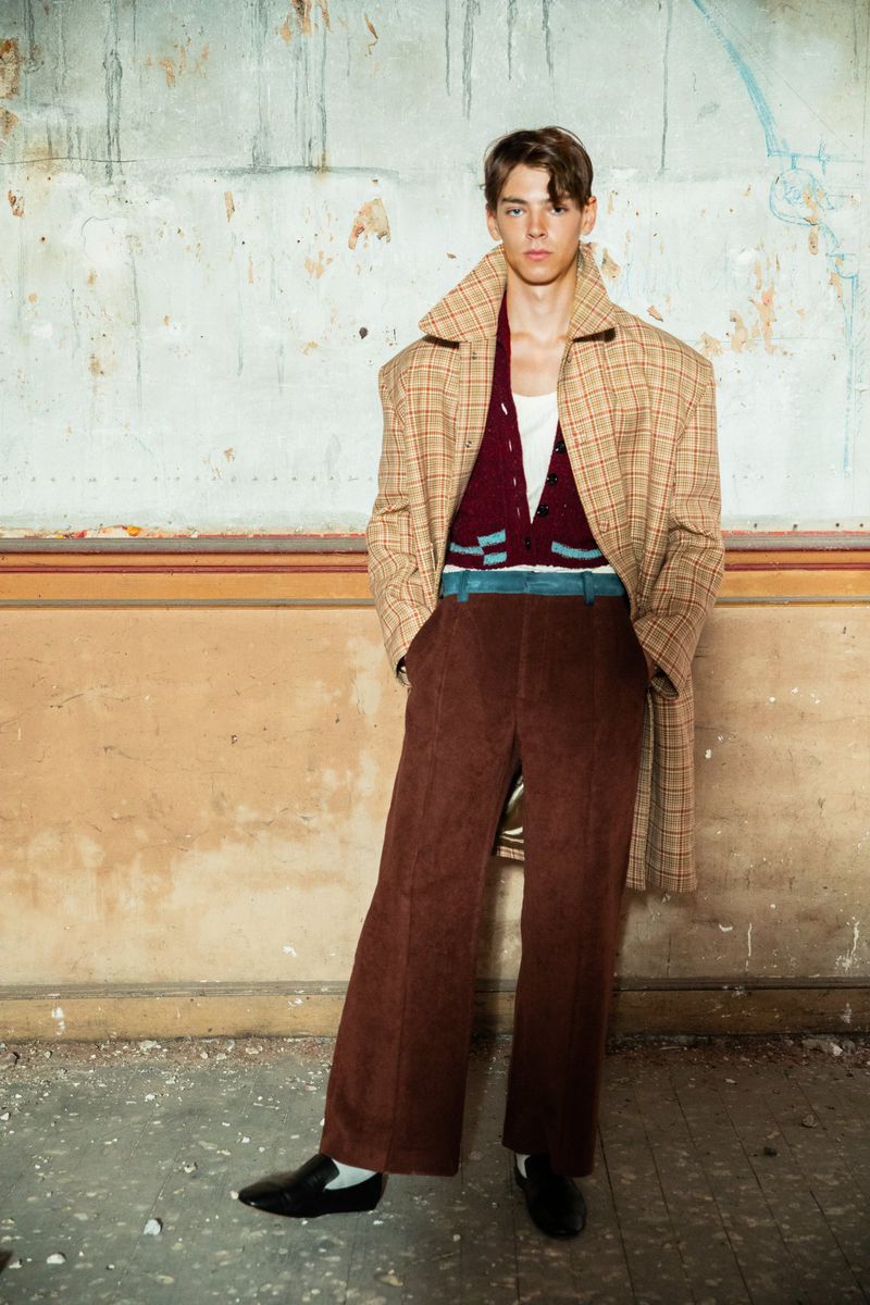 Young man in oversized coat and corduroy pants poses against a weathered wall, showcasing fashion apparel in a medium shot indoors.