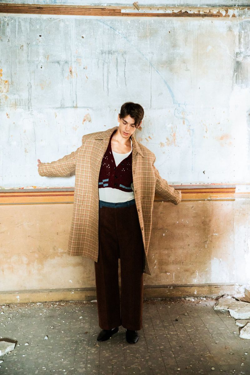 Young adult poses in oversized coat with maroon sweater vest, baggy brown pants, and black shoes against distressed wall.