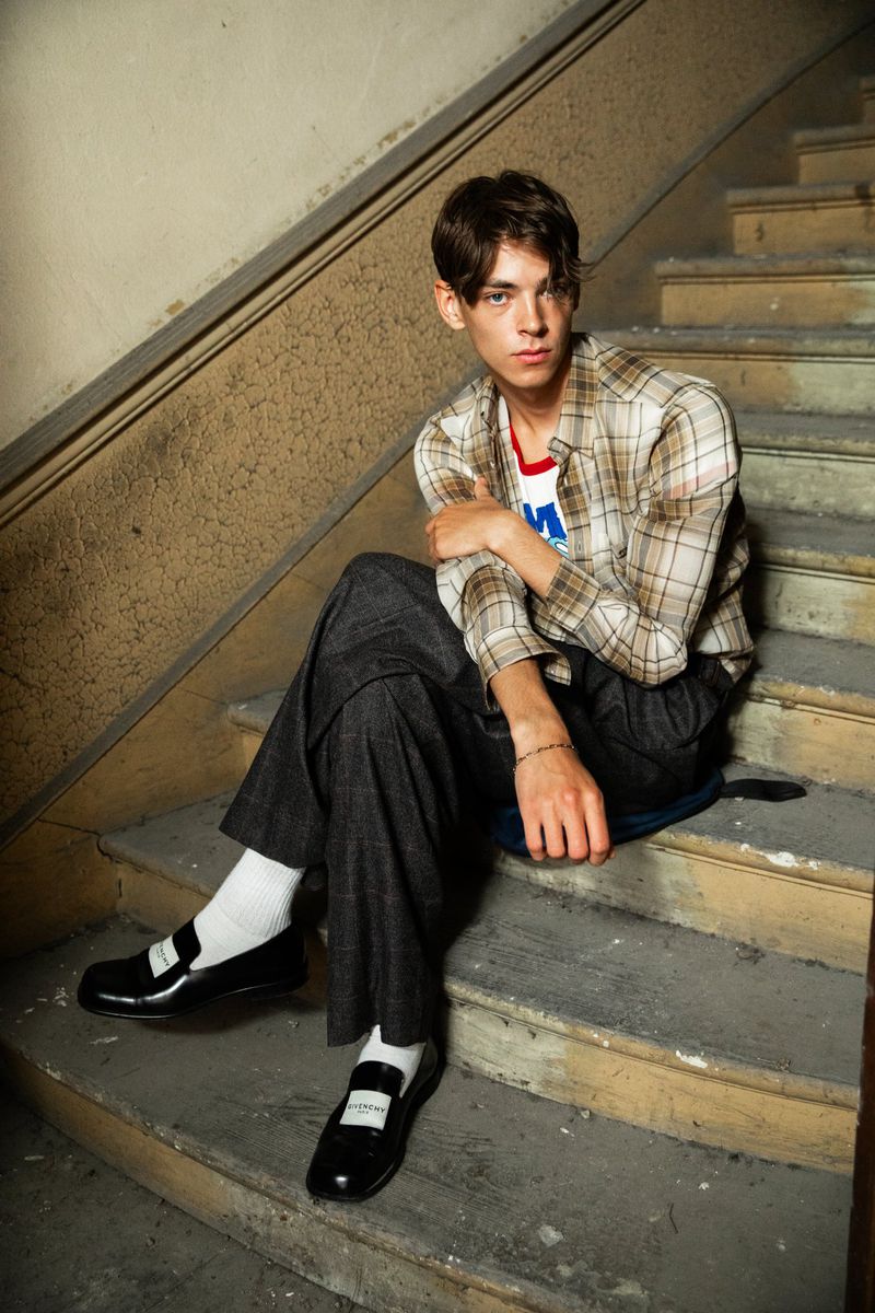 Young adult male poses sitting on stairs wearing a plaid shirt, dark pants, and black loafers with white socks indoors.