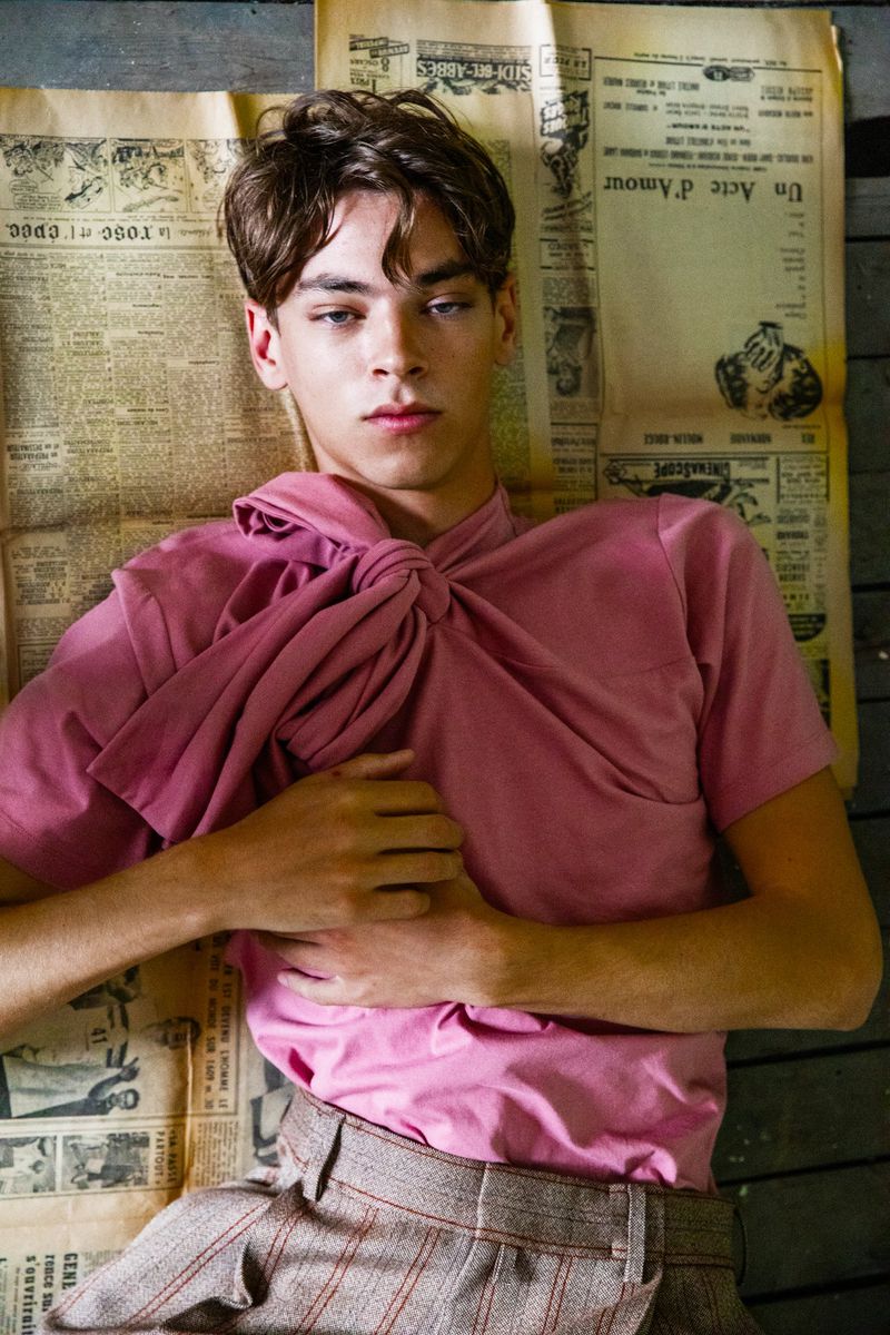 Young adult with brown hair lies on newspapers in a pink shirt and pinstripe pants looking into the camera in an indoor setting.
