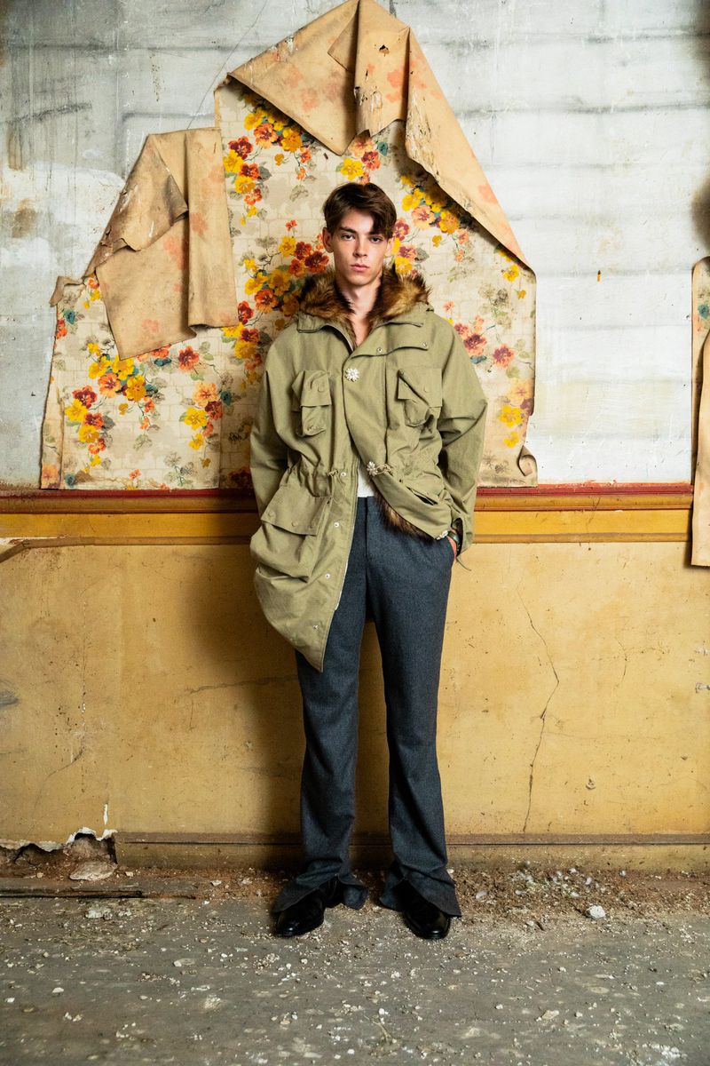 Full-length shot of a young adult male model wearing a parka, dress pants, and dress shoes in a distressed indoor location.