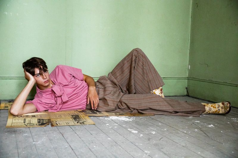 Young adult male lying on newspaper in pink shirt and brown pants in a photo studio setting with light green wall in background.