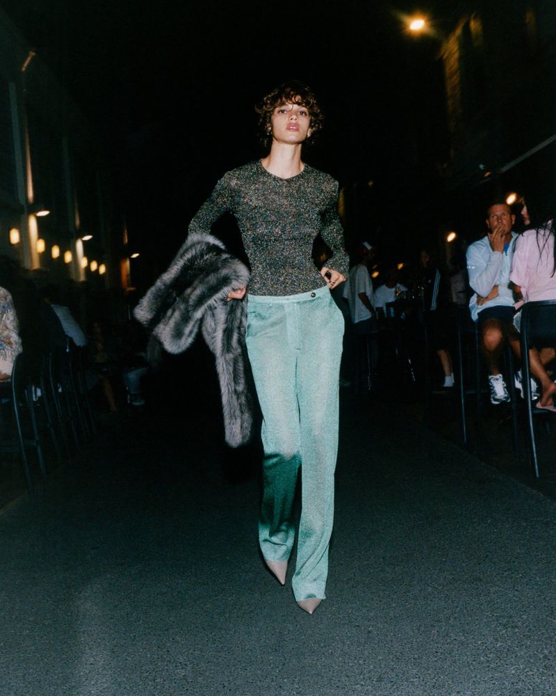 A person walks a dark runway in turquoise pants, a sheer long-sleeve top, and a fur coat during a fashion show with audience members.