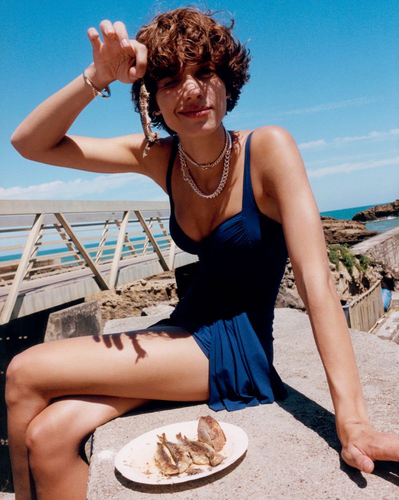 Woman in a swimsuit sits near the ocean, holding small cooked fish while posing for the camera on a sunny day with a plate of fish.