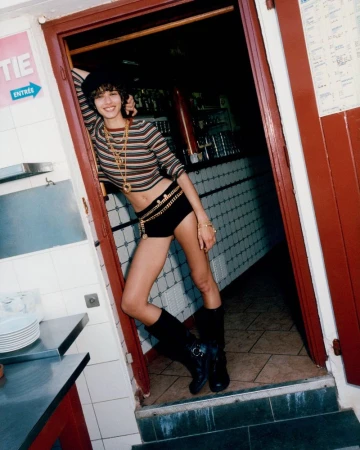A model poses indoors by a doorway wearing a striped top, black shorts with a chain belt, and black boots against a vintage-style backdrop.