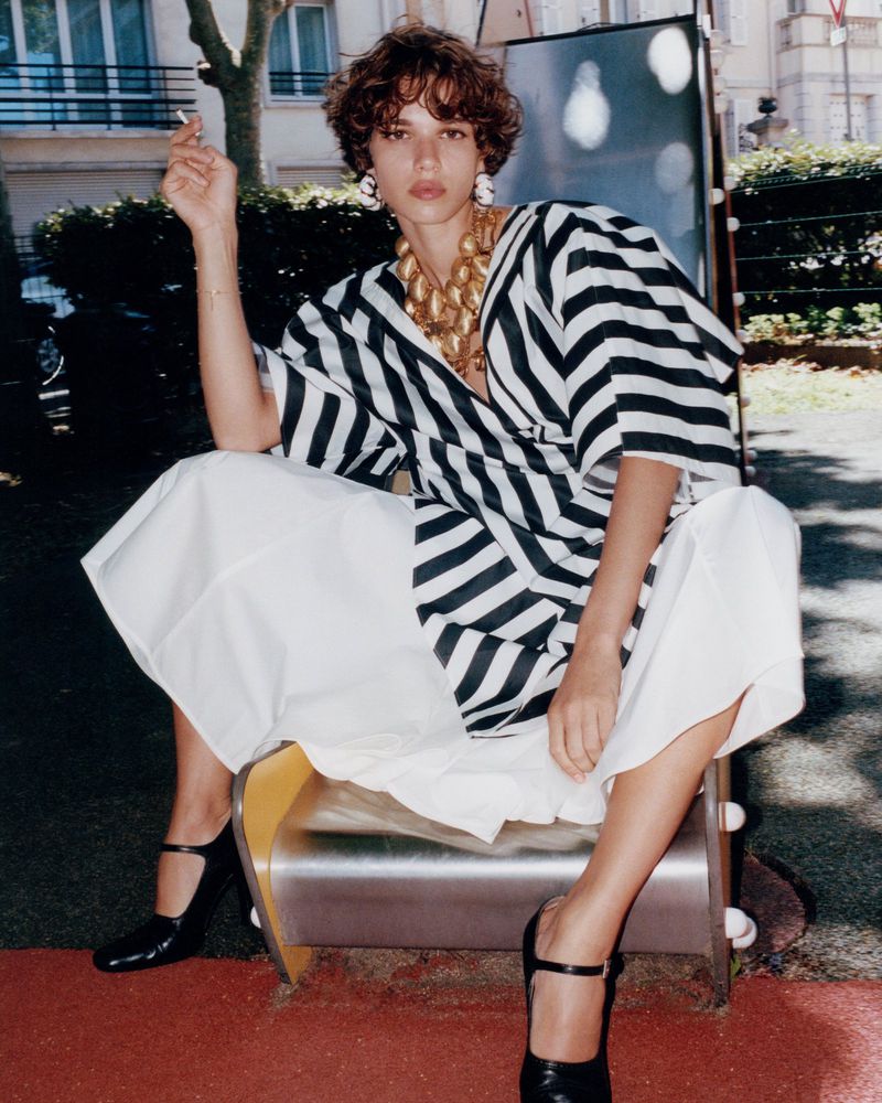 Fashion shot of a young adult female in a striped shirt and white skirt sitting on playground equipment, outdoors on a sunny day.