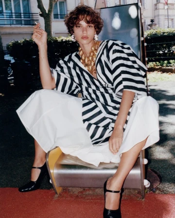 Fashion shot of a young adult female in a striped shirt and white skirt sitting on playground equipment, outdoors on a sunny day.