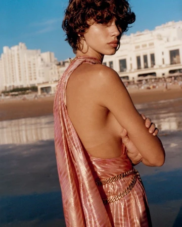 Mathilda Gvarliani posing on a sandy beach wearing a backless shimmering pink draped dress and gold chain belt.