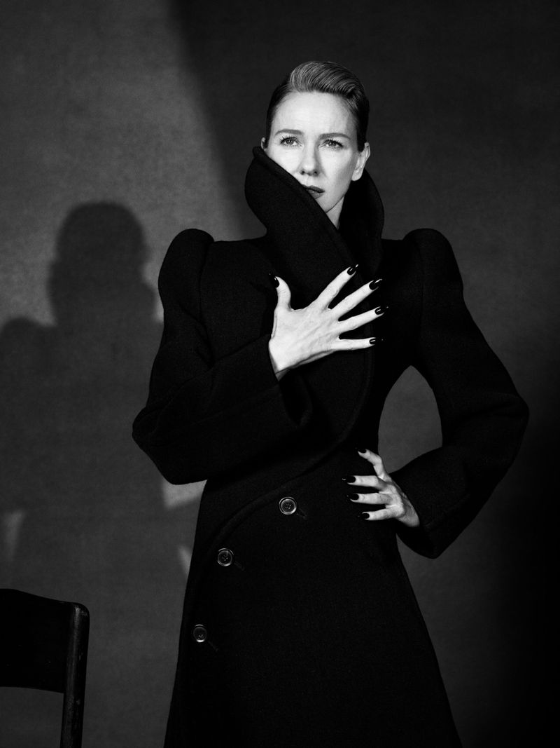 Naomi Watts in a black high-collar sculptural coat with structured shoulders, posing against a grey studio backdrop in black and white.
