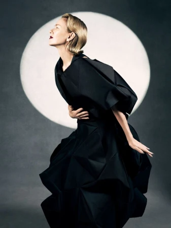 Elegant woman with short blonde hair posing in a structural black dress in front of a spotlight on a dark grey backdrop in a studio.