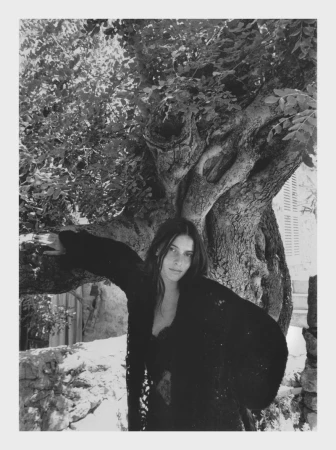 Monochrome portrait of a woman with long dark hair standing near a large tree in a rustic setting with stone structures.