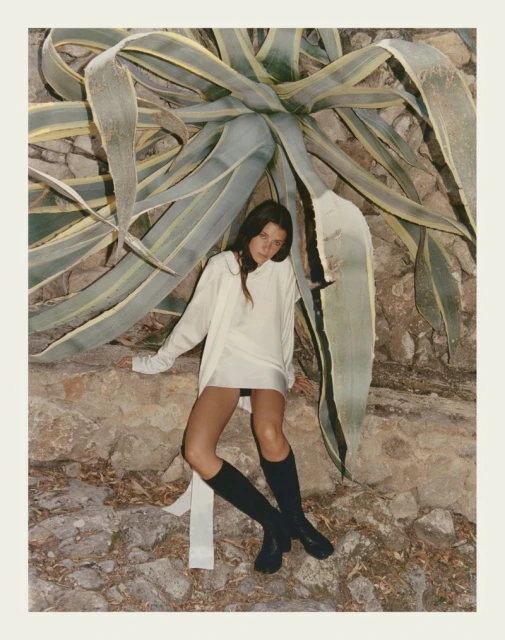 A brunette woman posing in a white mini dress and tall black boots, leaning against a stone wall with a large agave plant.