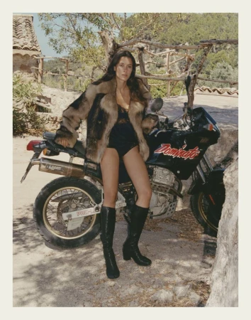 A woman in a fur coat and boots poses next to a black motorcycle on a sunny day in a rural setting, creating a fashion-forward outdoor shot.