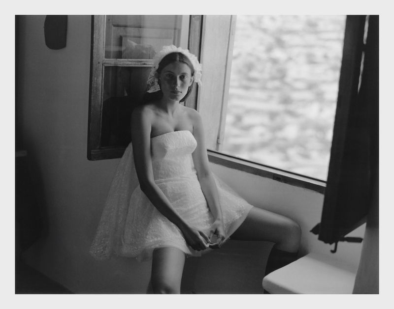 Woman in a white strapless dress and veil sits in front of a window in a black and white fashion photograph. 