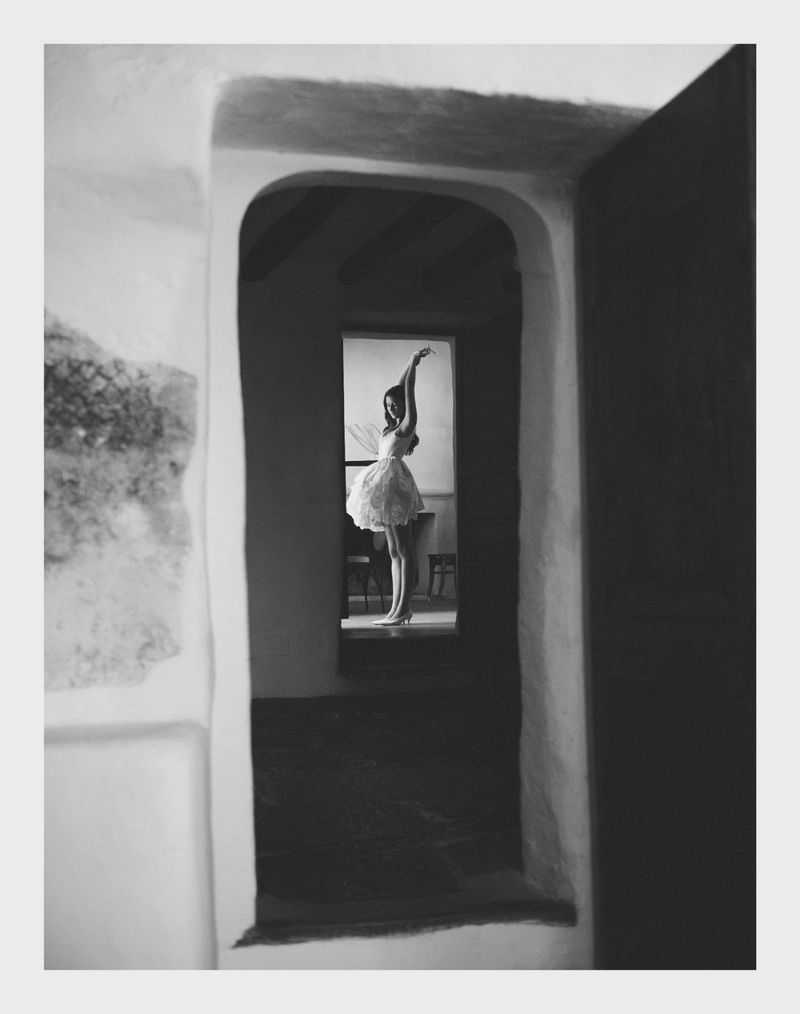 Monochrome shot of a woman in a white dress standing in a doorway with raised arms, viewed through multiple archways in an interior.