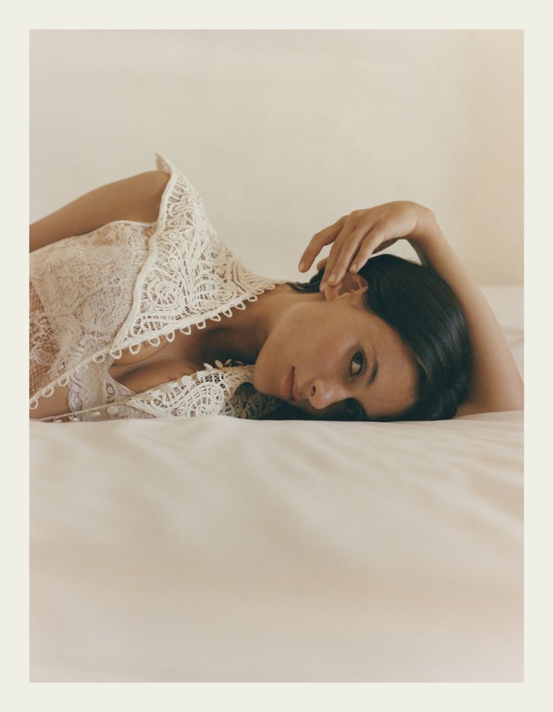 Adult female in lace top lying on white bed. Portrait-style shot with natural lighting on neutral background. Fashion, beauty, and lifestyle theme.