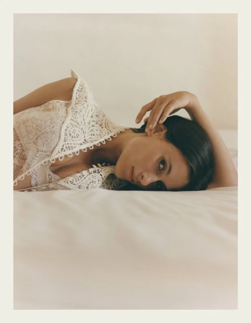 Adult female in lace top lying on white bed. Portrait-style shot with natural lighting on neutral background. Fashion, beauty, and lifestyle theme.