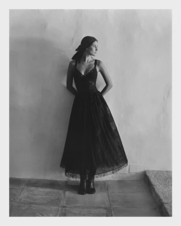 Woman in black dress and headscarf leans against a weathered wall on a tiled floor in this grayscale fashion portrait.