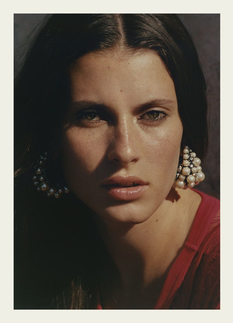 Close-up portrait of a beautiful woman with dark hair and green eyes wearing pearl earrings and a red top in soft, diffused light.