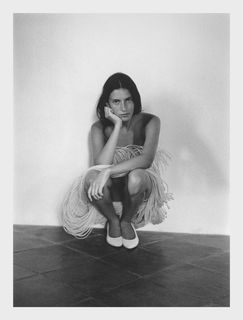 Monochrome portrait of a young woman squatting indoors, partially wrapped in beaded strings and wearing white shoes, looking at the camera.