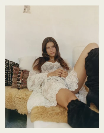 A young woman in a white lace dress and black high-heeled boots reclines on a tan upholstered surface in an indoor setting.