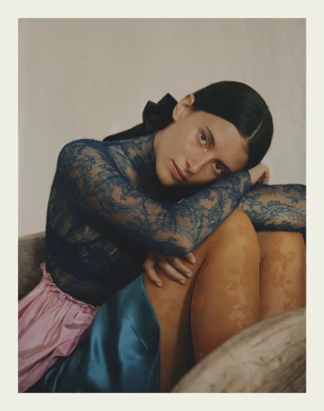 A young woman with dark hair wearing a blue lace top, pink skirt, and blue skirt poses indoors with soft, diffused lighting and a cream background.