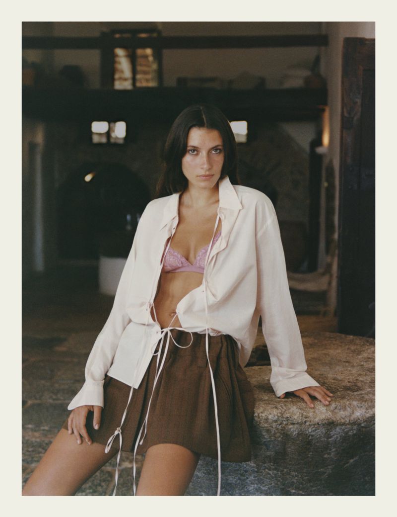 Brunette woman poses indoors wearing an unbuttoned white shirt and a brown skirt while leaning against a stone structure.