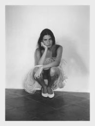 Model Lennon Sorrenti crouching against a wall wearing a looped cord garment and white flat shoes in a black and white portrait.