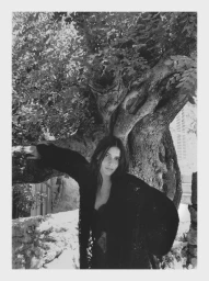 Model Lennon Sorrenti poses leaning against a large gnarled tree in a black outfit in a black and white editorial photograph.