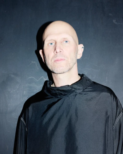 Headshot of a bald, adult male with light eyes wearing a dark top against a textured dark grey background in a simple studio setting.