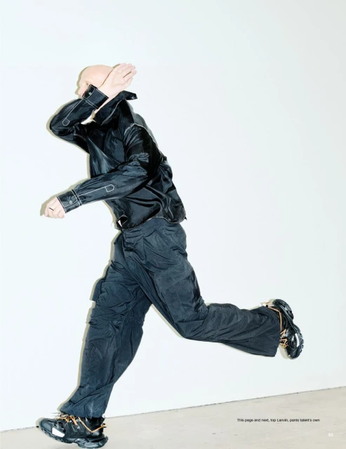 A bald adult male in a black jacket and pants is running across a light background. He has his hands up near his face. 