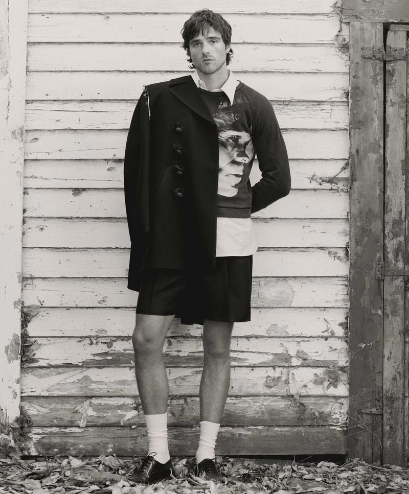 Monochrome full shot of adult man in patterned sweater, coat, shorts, socks, and loafers, standing against weathered wood background.