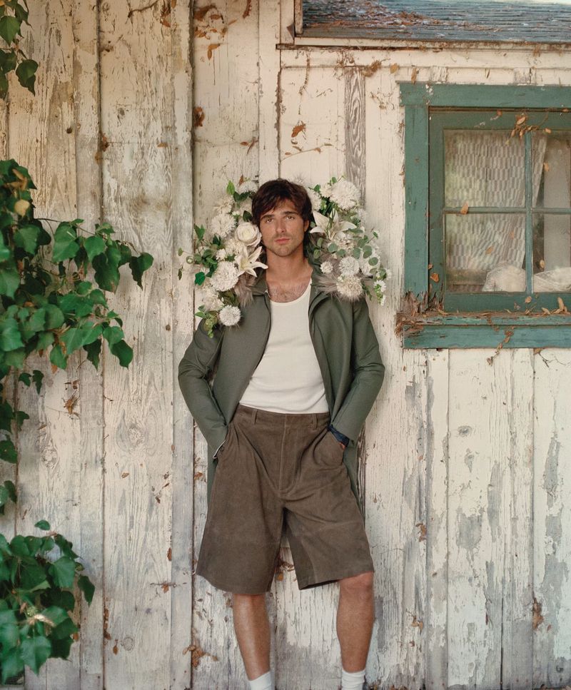 Portrait of a man wearing a jacket and shorts, standing against a weathered wooden wall, adorned with white flowers and greenery.