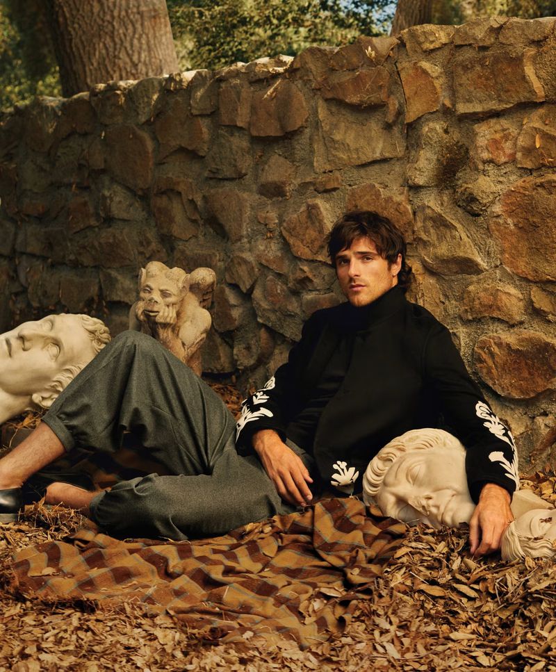 Adult male model in casual fashion, posed outdoors with sculpture busts and a stone wall in background. Fashion editorial shot.