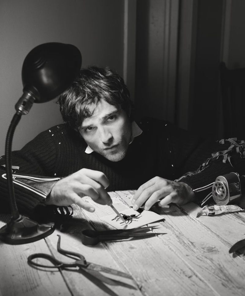 A man is hunched over a wooden table examining a spider with a tool under the light of a lamp, wearing a dark sweater, in a dim room.