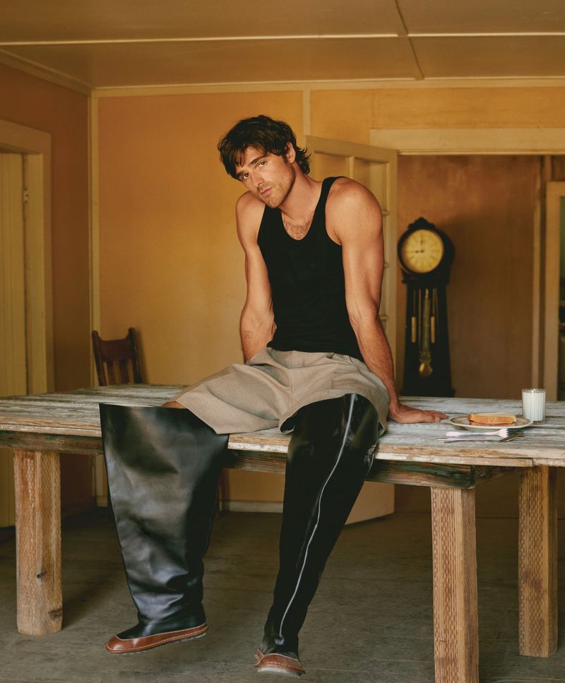 Man in black tank top and oversized leather boots sits on a wooden table indoors with vintage clock and plate of food in the background.
